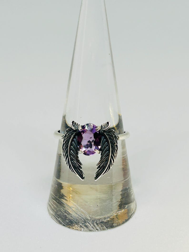 May include: A silver ring featuring a purple oval gemstone, flanked by two detailed feather designs. The ring is displayed on a clear, conical stand against a white background. The ring's design is intricate and elegant.