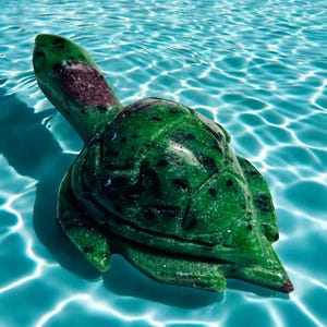 May include: A carved green stone turtle figurine with dark spots and a maroon patch on its head. The turtle is floating in clear blue water, creating a serene and naturalistic scene.