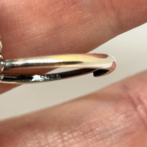 May include: Close-up of a silver ring with a curved design. The ring is highly polished, reflecting light. The band has a small marking. The background is a blurred skin tone.