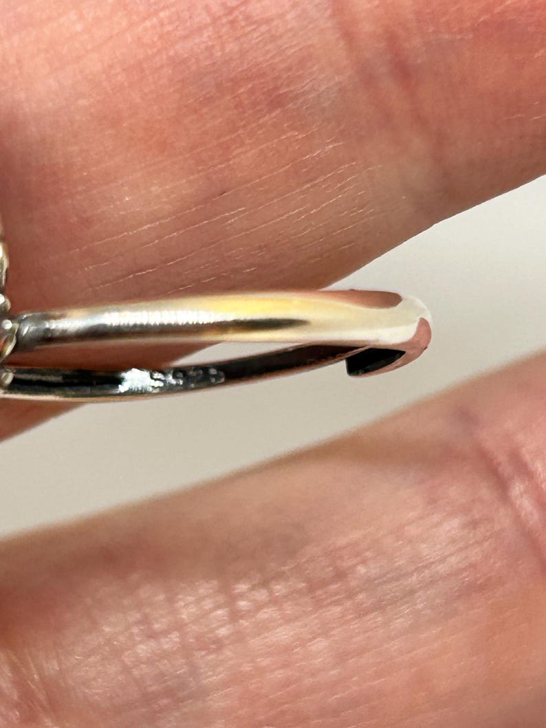May include: Close-up of a silver ring with a curved design. The ring is highly polished, reflecting light. The band has a small marking. The background is a blurred skin tone.