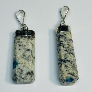 Może przedstawiać: Dwa wisiorki z kamienia K2 ze srebrnymi metalowymi elementami. Prostokątne wisiorki mają polerowane wykończenie, prezentując mieszankę kolorów białego, czarnego i niebieskiego. Przeznaczone do tworzenia biżuterii.