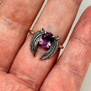 May include: A rose gold ring featuring a large oval-cut purple gemstone, flanked by detailed, dark silver feather accents. The ring is displayed on a hand, showcasing the intricate design and color contrast.