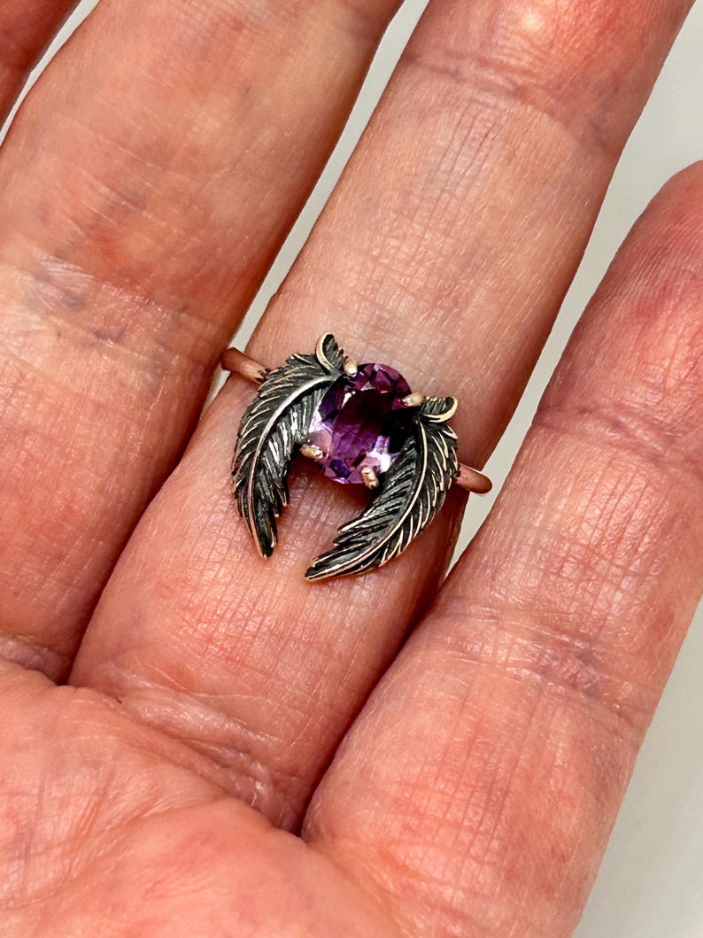 May include: A rose gold ring featuring a large oval-cut purple gemstone, flanked by detailed, dark silver feather accents. The ring is displayed on a hand, showcasing the intricate design and color contrast.