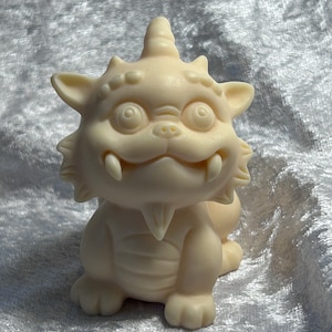 May include: A white ceramic figurine of a mythical creature with a horn on its head, large eyes, and a toothy grin. The creature is sitting on its haunches with its paws tucked under its body.
