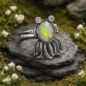 May include: A silver ring with an oval opal gemstone displaying iridescent colours. The ring has a distinctive design, with two small clear stones and an octopus-inspired base. The ring is presented on a dark grey stone.