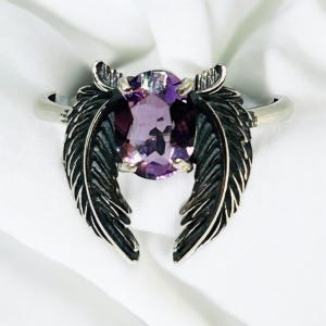 May include: A silver ring featuring a large, oval-cut purple gemstone. The ring's design includes two stylized feather accents on either side of the gemstone, creating a unique and elegant look. The ring is set against a white background.