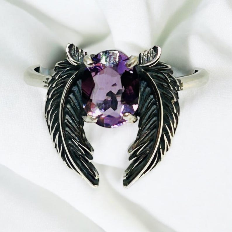 May include: A silver ring featuring a large, oval-cut purple gemstone. The ring's design includes two stylized feather accents on either side of the gemstone, creating a unique and elegant look. The ring is set against a white background.