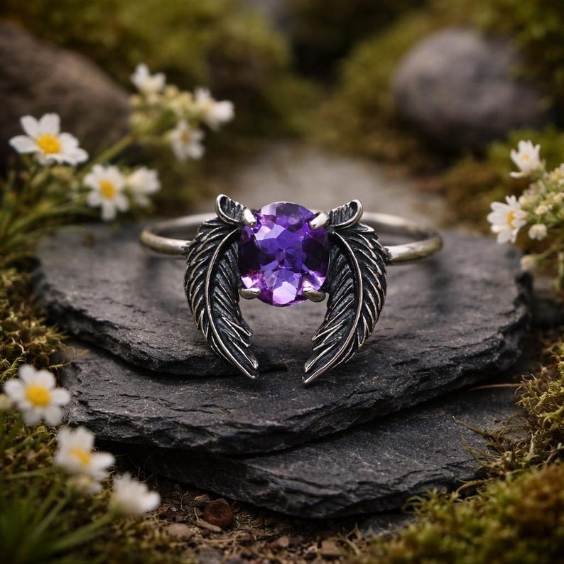 May include: A silver ring featuring a large, oval-cut purple gemstone, flanked by detailed feather designs. The ring is set on a dark gray stone surface, with a blurred background of greenery and small white flowers.