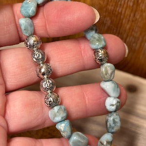 May include: A beaded bracelet featuring light blue and white larimar stones interspersed with silver-colored, patterned beads. The bracelet is held up, showcasing the natural variations in the stones and the intricate details of the metal beads.