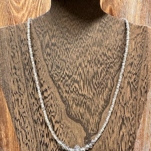 May include: A delicate silver necklace with small, clear beads. The necklace features a central pendant with a cluster of silver beads and a small, round, pink stone. The necklace is displayed on a wooden bust.