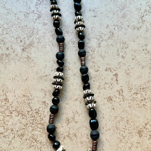 May include: A black beaded necklace with alternating sections of black and white beads. The necklace features copper-colored spacers and a variety of bead shapes, creating a unique and textured design. The necklace is displayed on a neutral-toned surface.