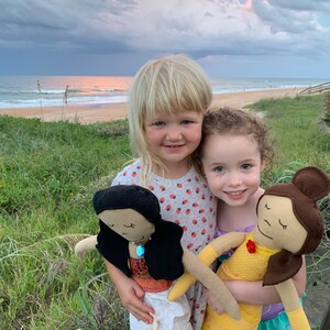 Peut inclure: Deux jeunes filles tiennent des poup&eacute;es faites &agrave; la main. Une poup&eacute;e a de longs cheveux noirs et porte une robe blanche. L'autre poup&eacute;e a les cheveux bruns et porte une robe jaune. Les filles sourient et regardent la cam&eacute;ra.