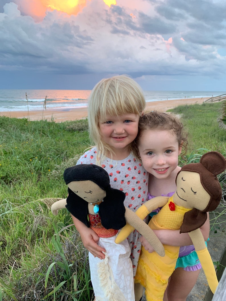Peut inclure: Deux jeunes filles tiennent des poup&eacute;es faites &agrave; la main. Une poup&eacute;e a de longs cheveux noirs et porte une robe blanche. L'autre poup&eacute;e a les cheveux bruns et porte une robe jaune. Les filles sourient et regardent la cam&eacute;ra.