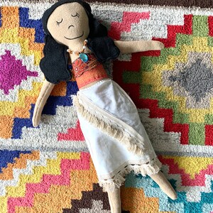 Peut inclure: Une poup&eacute;e faite &agrave; la main avec de longs cheveux noirs, portant une robe blanche avec un haut brun et orange et un collier bleu. La poup&eacute;e est allong&eacute;e sur un tapis color&eacute; &agrave; motifs g&eacute;om&eacute;triques.