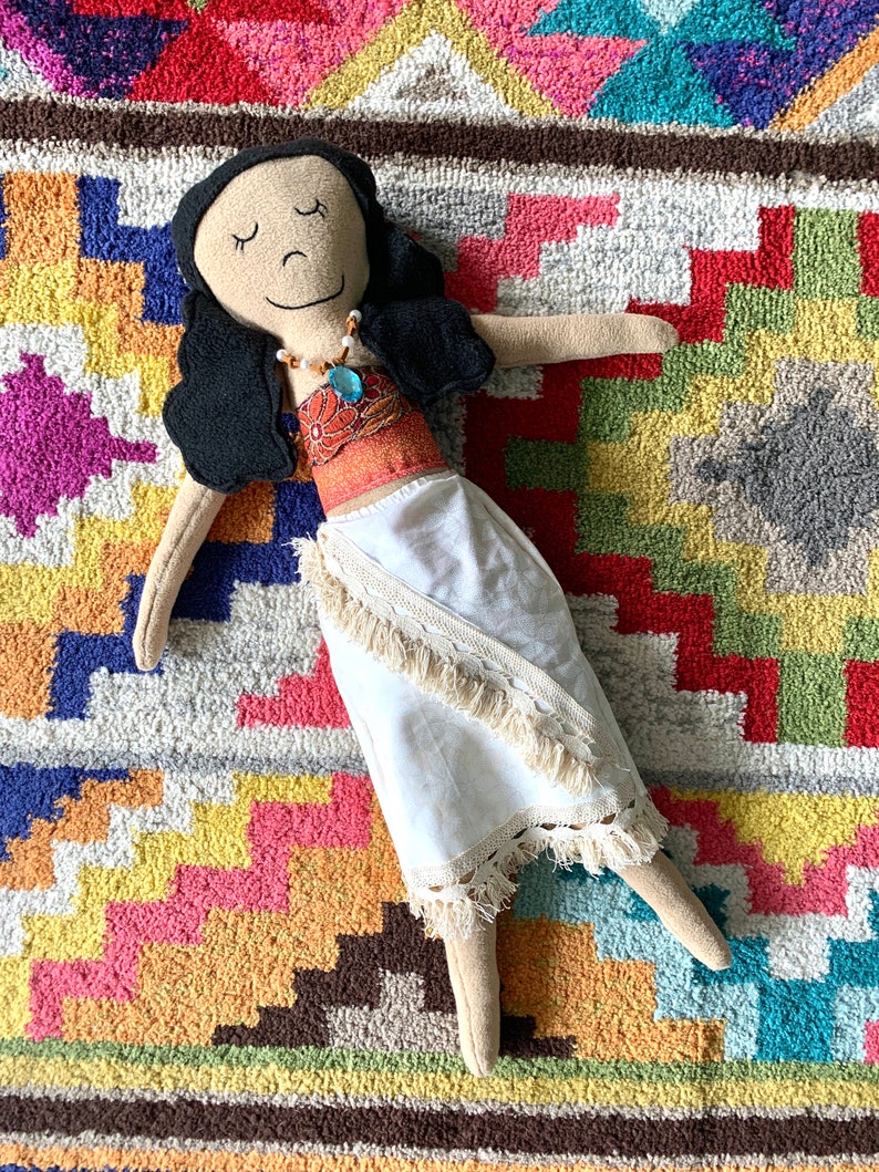 Peut inclure: Une poup&eacute;e faite &agrave; la main avec de longs cheveux noirs, portant une robe blanche avec un haut brun et orange et un collier bleu. La poup&eacute;e est allong&eacute;e sur un tapis color&eacute; &agrave; motifs g&eacute;om&eacute;triques.
