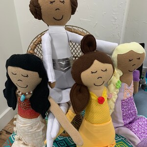 Peut inclure: Quatre poup&eacute;es en tissu faites &agrave; la main, chacune avec un th&egrave;me de princesse diff&eacute;rent. Les poup&eacute;es sont assises sur une chaise en osier et un tapis &agrave; motifs. Les poup&eacute;es portent toutes des robes et ont les cheveux bruns.
