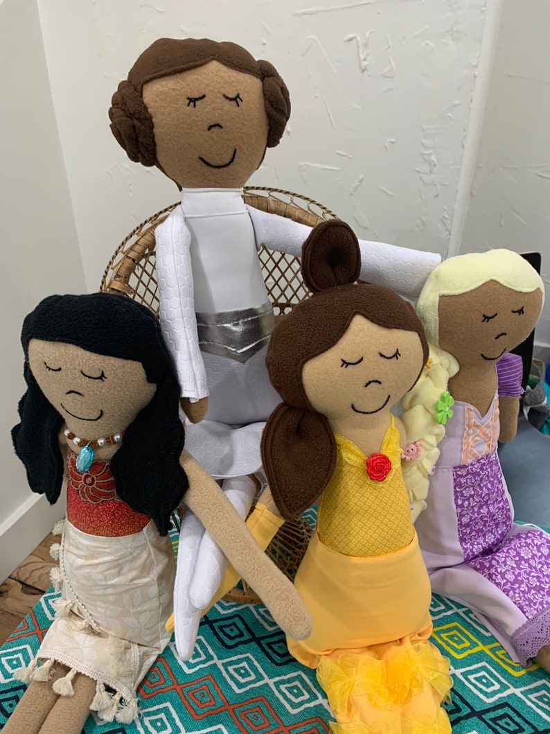 Peut inclure: Quatre poup&eacute;es en tissu faites &agrave; la main, chacune avec un th&egrave;me de princesse diff&eacute;rent. Les poup&eacute;es sont assises sur une chaise en osier et un tapis &agrave; motifs. Les poup&eacute;es portent toutes des robes et ont les cheveux bruns.