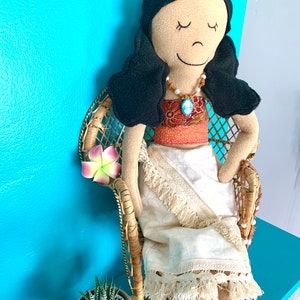 Peut inclure: Une poup&eacute;e faite &agrave; la main avec des cheveux noirs et un visage brun portant une robe blanche &agrave; franges et un collier brun et bleu. La poup&eacute;e est assise dans un fauteuil en osier avec une fleur rose.