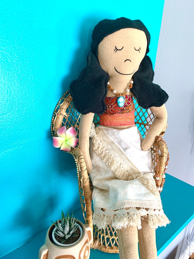 Peut inclure: Une poup&eacute;e faite &agrave; la main avec des cheveux noirs et un visage brun portant une robe blanche &agrave; franges et un collier brun et bleu. La poup&eacute;e est assise dans un fauteuil en osier avec une fleur rose.