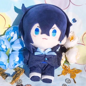 Puede incluir: Muñeco de peluche de un personaje con cabello azul oscuro y un traje a juego, con una pajarita azul claro. El muñeco tiene ojos azules bordados y una boca pequeña cosida. El muñeco está rodeado de flores azules y blancas.