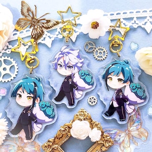 Op de afbeelding: Drie anime-personagecharms met gouden sleutelhangers. De charms zijn van helder acryl met kleurrijke illustraties van personages met blauw, wit en groen haar. Elke charm heeft een witte achtergrond met een bloemmotief.