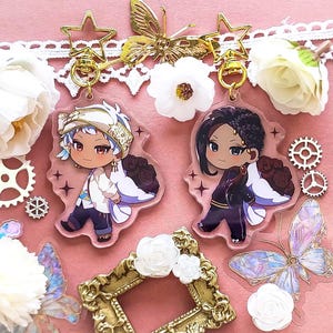 Op de afbeelding: Twee anime-achtige karaktercharms met gouden sleutelhangers. De charms zijn van helder acryl met zwart-wit illustraties van personages met zwart en wit haar. De personages dragen uitgebreide outfits en hebben grote ogen. De charms zijn omringd door witte bloemen en vlinders.
