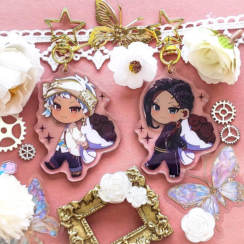 Op de afbeelding: Twee anime-achtige karaktercharms met gouden sleutelhangers. De charms zijn van helder acryl met zwart-wit illustraties van personages met zwart en wit haar. De personages dragen uitgebreide outfits en hebben grote ogen. De charms zijn omringd door witte bloemen en vlinders.