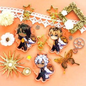 Op de afbeelding: Drie anime-achtige sleutelhangers met gouden ster-bedels. Elke sleutelhanger heeft een ander personage met een witte en gouden halo en een boeket gele bloemen. De personages zijn een man met groen haar, een man met blond haar en een man met wit haar.