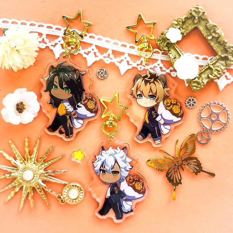 Op de afbeelding: Drie anime-achtige sleutelhangers met gouden ster-bedels. Elke sleutelhanger heeft een ander personage met een witte en gouden halo en een boeket gele bloemen. De personages zijn een man met groen haar, een man met blond haar en een man met wit haar.