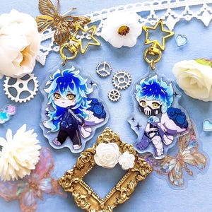 Op de afbeelding: Twee blauwe en witte anime-personagecharms met gouden stercharms en gouden kettingschakels. De charms bevinden zich op een lichtblauwe achtergrond met witte bloemen, gouden tandwielen en vlinders.