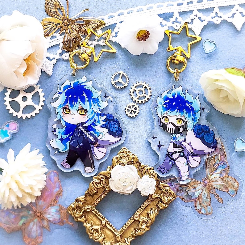 Op de afbeelding: Twee blauwe en witte anime-personagecharms met gouden stercharms en gouden kettingschakels. De charms bevinden zich op een lichtblauwe achtergrond met witte bloemen, gouden tandwielen en vlinders.