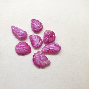 May include: Seven carved leaf-shaped gemstones in shades of pink and white are arranged on a light background. The gemstones have detailed veining and a polished finish, suggesting they are for jewelry making or decorative purposes.