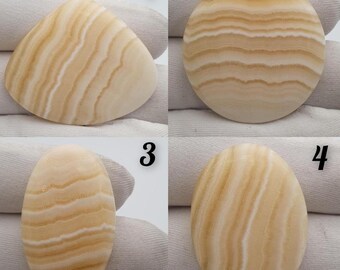May include: Four polished gemstones with a layered, banded pattern in shades of cream and tan. The shapes include a triangle, a circle, and two oval stones. The stones are displayed on a white surface.