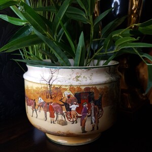 May include: A large, cream-coloured ceramic planter with a green rim. The planter features a hand-painted scene of a horse-drawn carriage with people in period clothing. The planter is filled with green leafy plants.