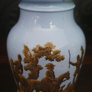 May include: A light blue ceramic vase with a gold design of people riding horses in a forest. The vase has a wide mouth and a rounded body.