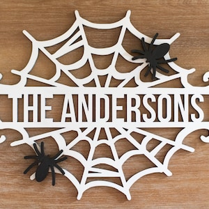 May include: White wooden Halloween sign shaped like a spider web with the text "The Andersons" in the center. Two black spiders are attached to the web.
