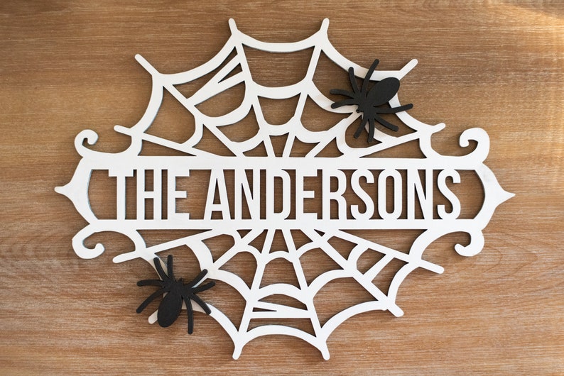 Puede incluir: Un letrero de Halloween de madera blanca con forma de telara&ntilde;a con el texto "The Andersons" en el centro. Dos ara&ntilde;as negras est&aacute;n unidas a la telara&ntilde;a.