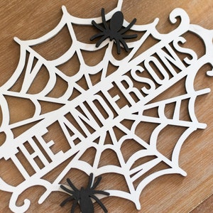 Puede incluir: Un letrero de Halloween de madera blanca con un dise&ntilde;o de telara&ntilde;a y el texto "The Andersons". Dos ara&ntilde;as negras est&aacute;n unidas a la telara&ntilde;a.