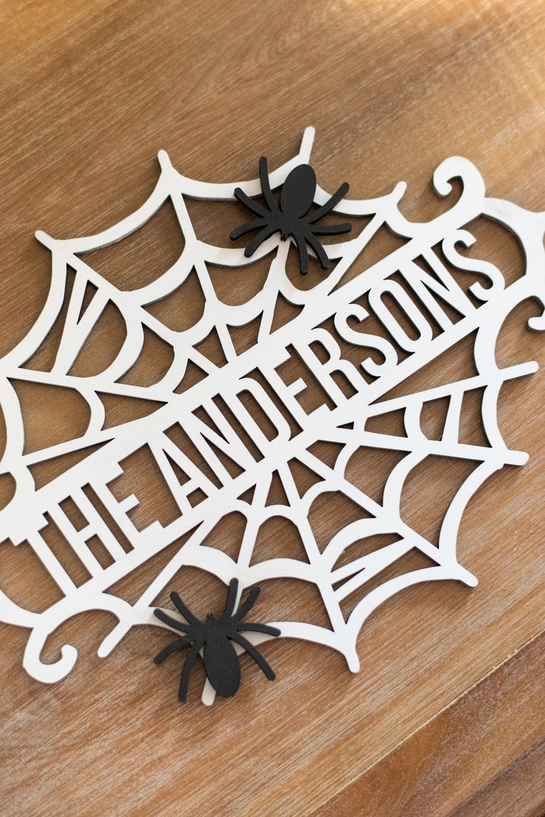 Puede incluir: Un letrero de Halloween de madera blanca con un dise&ntilde;o de telara&ntilde;a y el texto "The Andersons". Dos ara&ntilde;as negras est&aacute;n unidas a la telara&ntilde;a.
