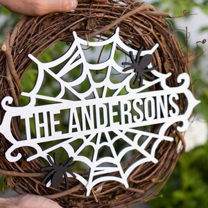 Puede incluir: Una corona de Halloween de madera blanca con un dise&ntilde;o de telara&ntilde;a y el texto "The Andersons". La corona est&aacute; decorada con ara&ntilde;as negras.