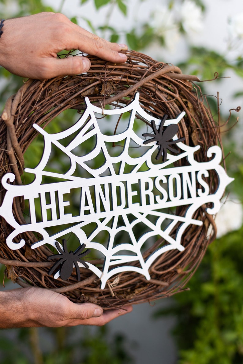 Puede incluir: Una corona de Halloween de madera blanca con un dise&ntilde;o de telara&ntilde;a y el texto "The Andersons". La corona est&aacute; decorada con ara&ntilde;as negras.