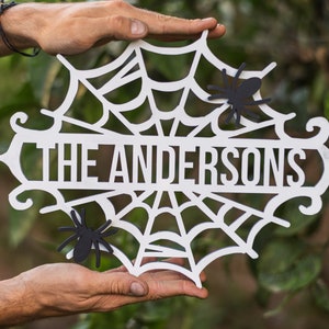 Puede incluir: Un letrero de madera blanco de Halloween con un dise&ntilde;o de telara&ntilde;a y el texto "THE ANDERSONS" en letras negras. Dos ara&ntilde;as de papel negras est&aacute;n pegadas al letrero.