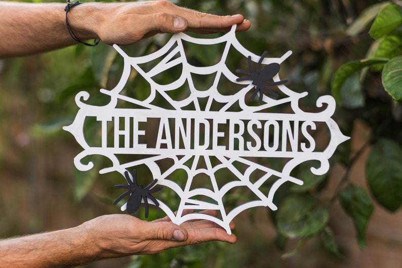 Puede incluir: Un letrero de madera blanco de Halloween con un dise&ntilde;o de telara&ntilde;a y el texto "THE ANDERSONS" en letras negras. Dos ara&ntilde;as de papel negras est&aacute;n pegadas al letrero.