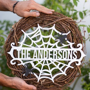 Puede incluir: Una corona de Halloween de madera blanca con un dise&ntilde;o de telara&ntilde;a y el texto "The Andersons". Dos ara&ntilde;as de papel negro est&aacute;n unidas a la corona.