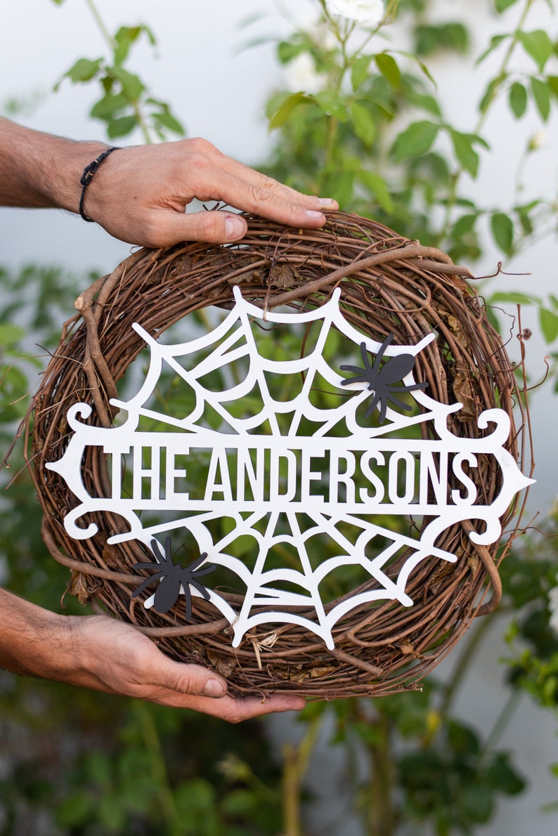 Puede incluir: Una corona de Halloween de madera blanca con un dise&ntilde;o de telara&ntilde;a y el texto "The Andersons". Dos ara&ntilde;as de papel negro est&aacute;n unidas a la corona.