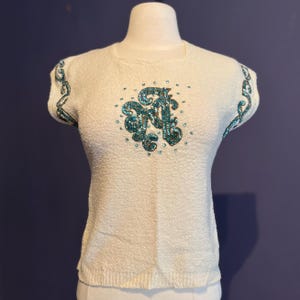 May include: A white knit sweater with short sleeves and a turquoise beaded design on the front. The design features a stylized letter "R" with swirls and embellishments.