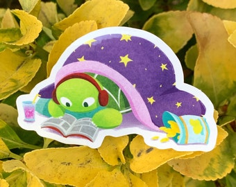 May include: A die-cut sticker showing a cartoon turtle under a purple blanket with yellow stars, wearing headphones and reading a book. A drink and a snack container are nearby. The turtle is green, and the blanket has a pink trim.