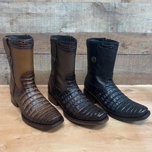 May include: Three leather cowboy boots in brown and black. The boots feature a textured, crocodile-like pattern and a square toe. Each boot has a decorative metal button. The boots are displayed on a wooden surface.