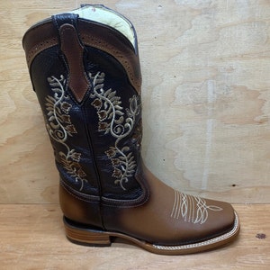 Op de afbeelding: Bruine leren cowboylaarzen met een vierkante neus en ingewikkelde bloemenborduursels in goudkleurig garen. De laarzen hebben een bruine leren schacht met een decoratief treklipje.