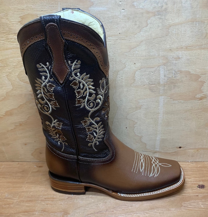 Op de afbeelding: Bruine leren cowboylaarzen met een vierkante neus en ingewikkelde bloemenborduursels in goudkleurig garen. De laarzen hebben een bruine leren schacht met een decoratief treklipje.